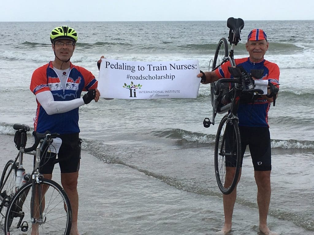 Jeff on the beach with fellow bike rider holding "Pedaling to Train Nurses" sign
